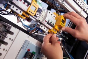 Home Service Blogs | Grand Bay Electrical Hands Installing A Yellow Circuit Breaker In An Electrical Panel With Wires And Switches Visible.