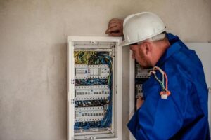 Home Service Blogs | Grand Bay Electrical Electrician In Blue Overalls And Helmet Examines A Control Panel With Wires And Switches.