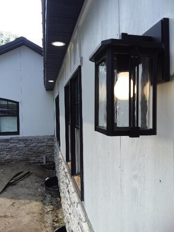 Enjoy Extra Light On The Shorter Days This Winter | Grand Bay Electrical Outdoor Wall-Mounted Lantern With A Glowing Bulb Is Attached To A Beige Wall. The Building Has Stone Accents And Multiple Windows Along The Side.
