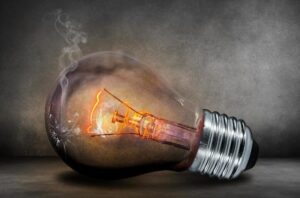 Home Service Blogs | Grand Bay Electrical A Light Bulb Emits Smoke With Its Filament Glowing Inside, Set Against A Textured Gray Background.