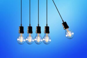 Home Service Blogs | Grand Bay Electrical Five Hanging Light Bulbs Against A Blue Background, With One Bulb Swinging Outward.