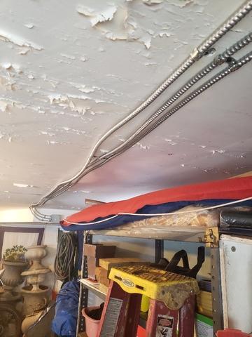Ceiling With Peeling Paint And Exposed Electrical Conduits, Suggesting Someone Was Running Power To A Grand Rapids Garage. Shelving Below Holds Various Items, Including A Ladder And Tarp.