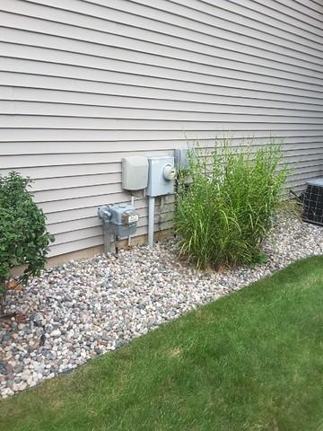 Side Of A House With A Beige Siding, Featuring Utility Meters, Pipes, And A Newly Completed Generac Generator Installation. Decorative Pebbles And Ornamental Grass Are Neatly Arranged Along The Foundation.