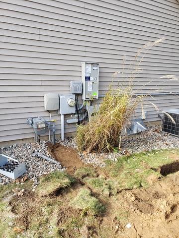 A House Exterior With Utility Panels, A Gas Meter, And Overgrown Grass Against The Wall Showcases A Recent Generac Generator Installation, Surrounded By Small Rocks And Partially Dug Soil In The Foreground.