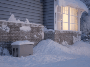 A Home With Snow-Covered Ground And Rooftop, Illuminated Window, And An Outdoor Generator Partially Covered In Snow Near The Brick Foundation.