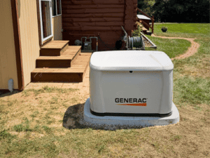A Generac Standby Generator Sits On A Concrete Pad Next To A House With Wooden Steps, Near A Garden Hose And A Grassy Yard.