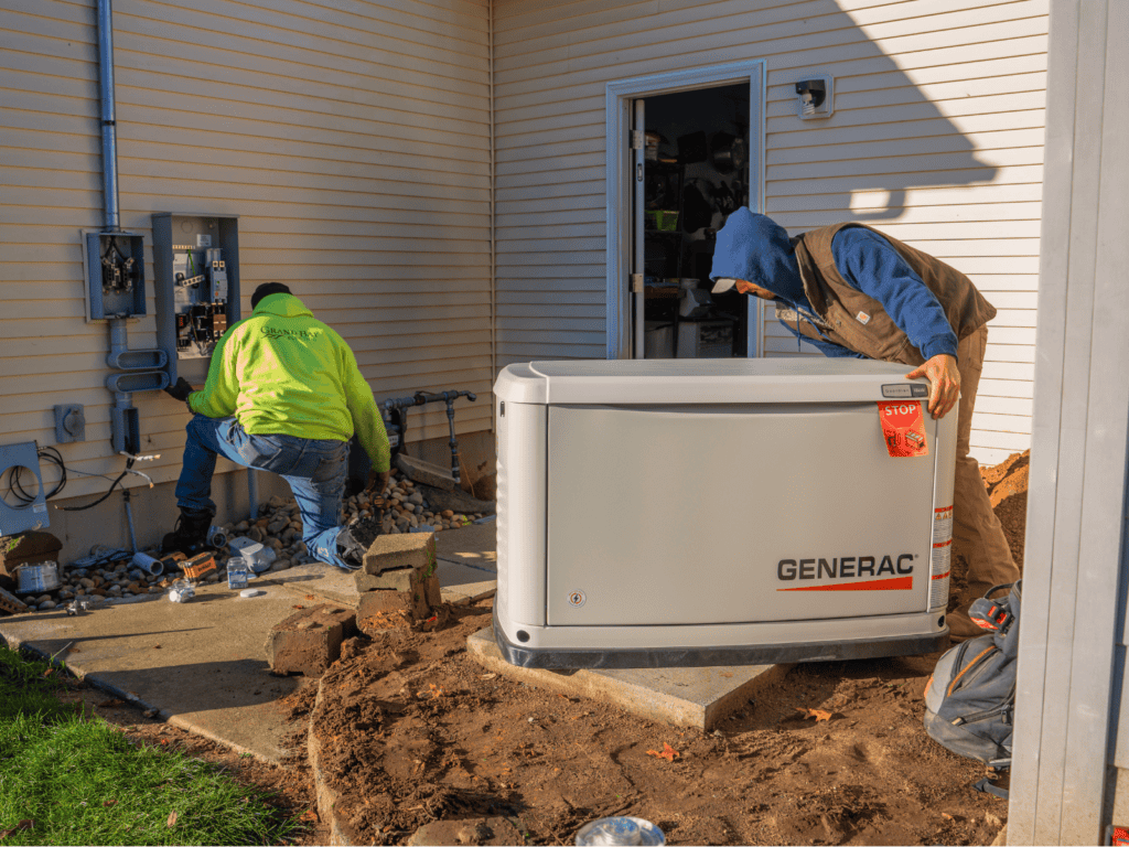 What To Expect During A Generator Installation | Grand Bay Electrical Two Workers Install A Generac Generator Outside A House; One Works At The Electrical Panel, The Other Adjusts The Generator On A Concrete Pad.